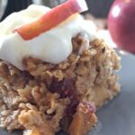 a square piece of apple cinnamon baked oatmeal with Cranberries and Pecanstopped with a dollup of Greek yogurt and a drizzle of honey sitting on a light gray plate